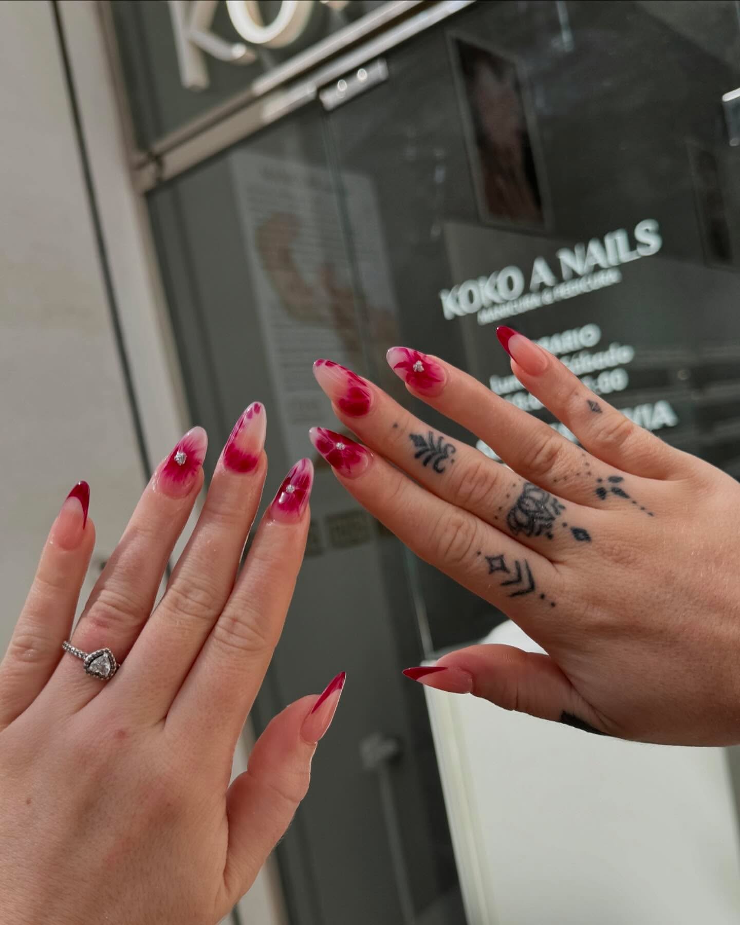 Dos manos con anillo y uñas almendradas en degradado rosa con flores rojas, frente al escaparate de Koko A Nails